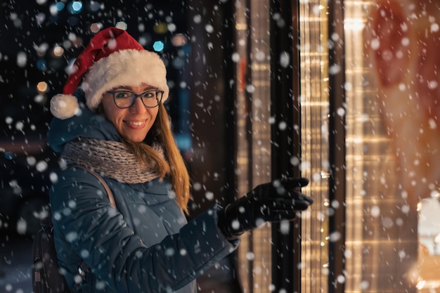 Hübsche Frau mit Weihnachtsmütze, die im beleuchteten Schaufenster schaut und träumt. Weihnachten präsentiert Feiertage oder Einkaufen zum Neujahrs- oder Weihnachtskonzept
