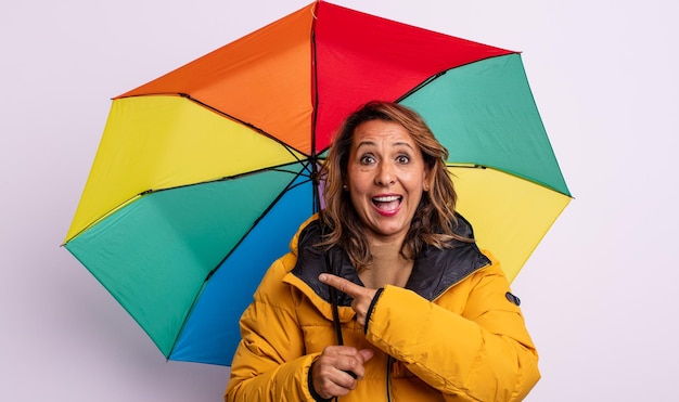 Hübsche Frau mittleren Alters, die aufgeregt und überrascht aussieht und auf das seitliche Regenschirmkonzept zeigt