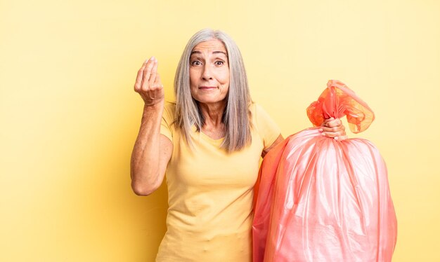 Hübsche Frau mittleren Alters, die Capice oder Geldgeste macht und Ihnen sagt, dass Sie bezahlen sollen. Müllbeutel aus Plastik