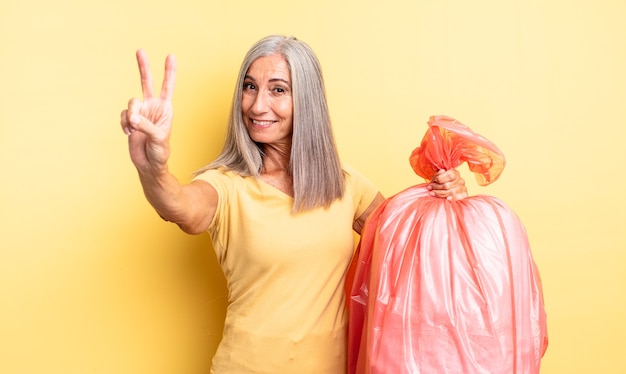 Hübsche Frau mittleren Alters, die lächelt und glücklich aussieht und Sieg oder Frieden gestikuliert. Müllbeutel aus Plastik