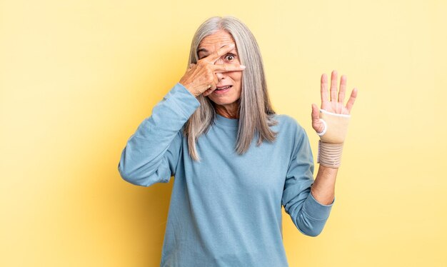 Hübsche Frau mittleren Alters, die schockiert, verängstigt oder verängstigt aussieht und das Gesicht mit Hand-Hand-Verband-Konzept bedeckt