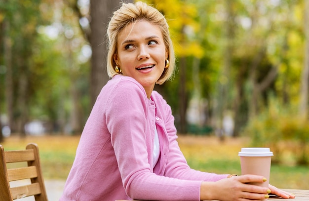 Hübsche Frau trinkt Kaffee auf der Terrasse Cafeteria im Park Blonde Frau in der Stadtstraße trinkt ein heißes Getränk Blick auf eine Seite Hübsches Mädchen mit einer Tasse Kaffee Kaffee für unterwegs