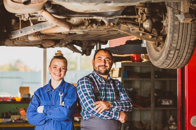 Hübsche Frau und gutaussehender Mann, beide Autoservice-Arbeiter, die Arbeitsoveralls tragen, stehen unter einem angehobenen Auto mit gekreuzten Händen und einem netten Lächeln, das in die Kamera schaut