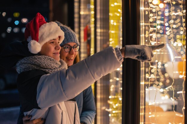 Hübsche Frau und ihr Sohn in Weihnachtsmütze schauen und träumen im beleuchteten Schaufenster