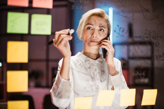 Foto hübsche geschäftsfrau, die im modernen büro auf glastafel schreibt und telefoniert