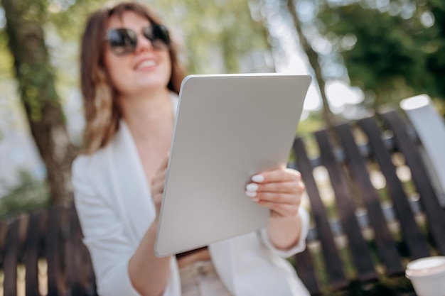 Hübsche Geschäftsfrau im weißen Anzug, die in der Parklandschaft der Stadt sitzt und an einem digitalen Tablet mit Elektroroller auf backgroundxA arbeitet