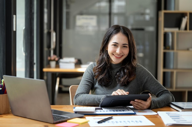 Hübsche Geschäftsfrau lächelt in die Kamera, während sie mit einem digitalen Tablet an ihrem Schreibtisch vor dem Computer sitzt
