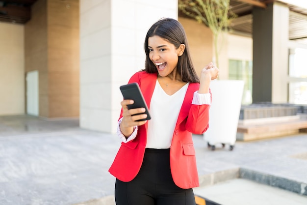 Hübsche Geschäftsfrau mit roter Jacke beim Lesen positiver Nachrichten auf dem Smartphone außerhalb des Büros