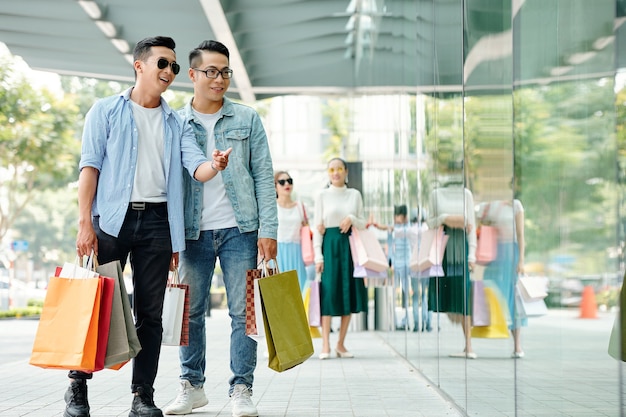 Hübsche glückliche junge asiatische Kerle, die zusammen einkaufen, wählen Turnschuhe im Schaufenster