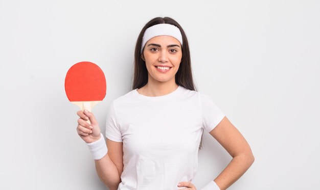 Hübsche hispanische Frau, die glücklich mit einer Hand auf der Hüfte und einem selbstbewussten Ping-Pong-Konzept lächelt