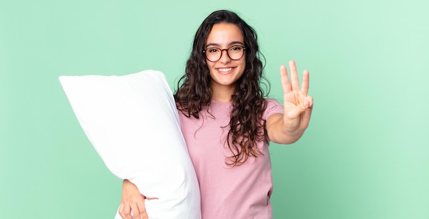 Hübsche hispanische Frau, die lächelt und freundlich aussieht, Nummer drei zeigt und Pyjamas mit einem Kissen trägt