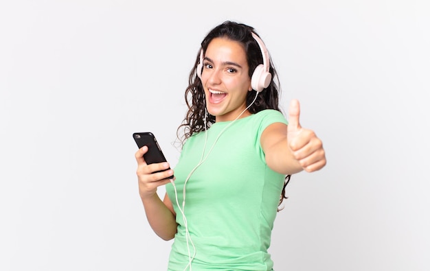 Hübsche hispanische Frau, die sich mit Kopfhörern und einem Smartphone stolz lächelt und positiv mit Daumen nach oben lächelt