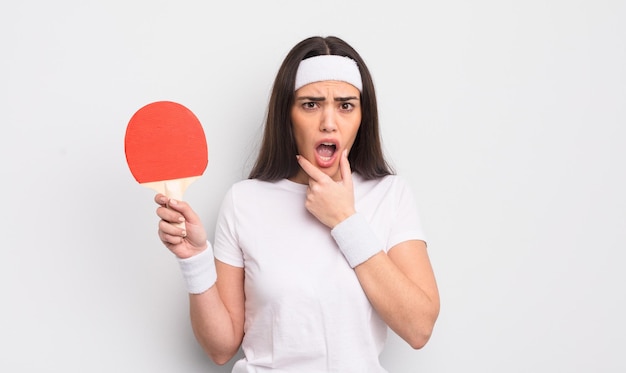 Hübsche hispanische Frau mit weit geöffnetem Mund und Augen und Hand auf dem Kinn-Ping-Pong-Konzept