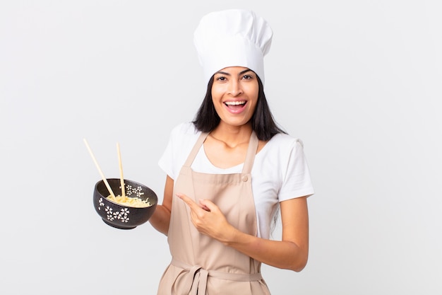 hübsche hispanische Kochfrau, die aufgeregt und überrascht aussieht, auf die Seite zeigt und eine Nudelschüssel hält