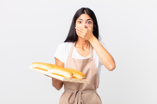 Hübsche hispanische Kochfrau, die den Mund mit den Händen mit einem schockierten bedeckt und ein Tablett mit Brotbrötchen hält