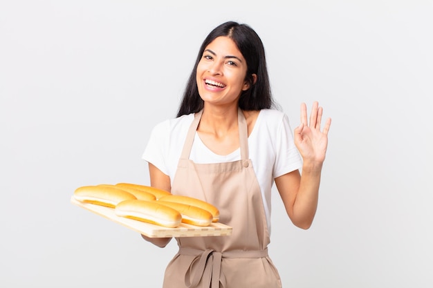 Hübsche hispanische Kochfrau, die glücklich lächelt, Hand winkt, Sie begrüßt und begrüßt und ein Tablett mit Brotbrötchen hält