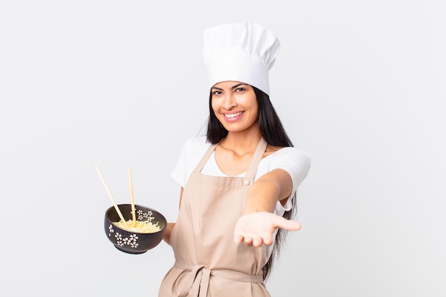 Hübsche hispanische Kochfrau, die glücklich mit freundlichem Lächeln lächelt und ein Konzept anbietet und zeigt und eine Nudelschüssel hält?