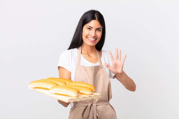 Hübsche hispanische Kochfrau, die lächelt und freundlich aussieht, Nummer fünf zeigt und ein Tablett mit Brotbrötchen hält?