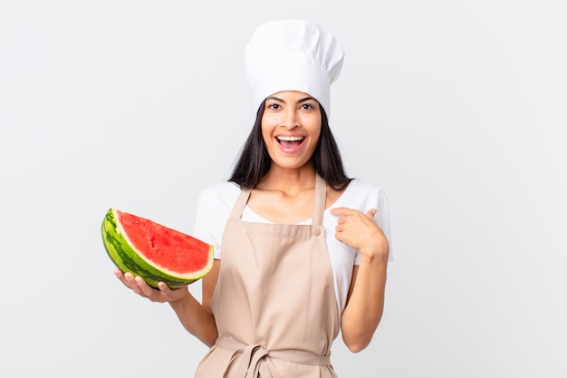 Hübsche hispanische Kochfrau, die sich glücklich fühlt und auf sich selbst zeigt, mit einer aufgeregten und hält eine Wassermelone