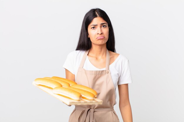 Hübsche hispanische Kochfrau, die sich traurig und weinerlich mit einem unglücklichen Blick fühlt und weint und ein Tablett mit Brotbrötchen hält