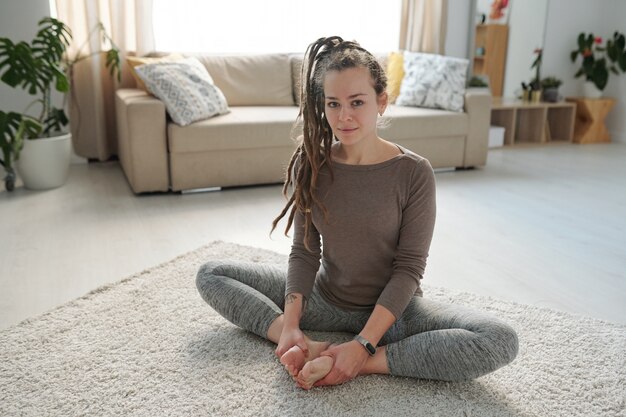 Hübsche junge aktive Frau, die ihre nackten Sohlen zusammensetzt, während sie während des Yoga auf dem Boden sitzen