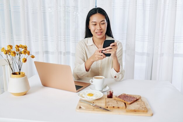 Hübsche junge Asiatin, die am Tisch sitzt, frühstückt und Nachrichten auf dem Smartphone und E-Mails auf dem Laptop überprüft