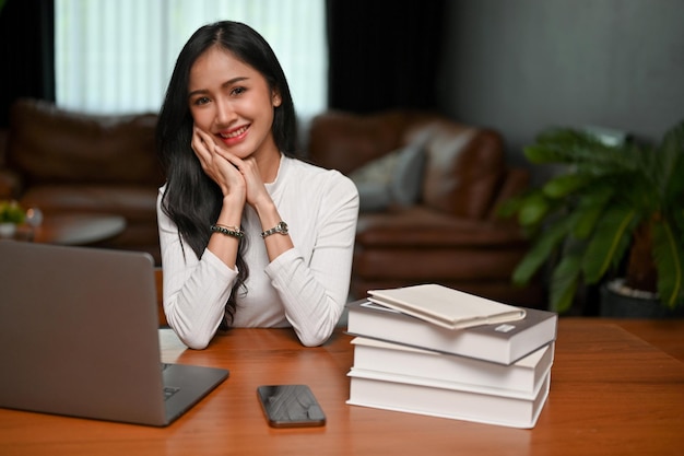 Hübsche junge asiatische Freiberuflerin oder Geschäftsfrau sitzt an ihrem Schreibtisch in ihrem Wohnzimmer