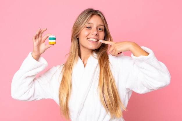 Hübsche junge blonde Frau, die Bademantel trägt und ein kosmetisches Produkt hält