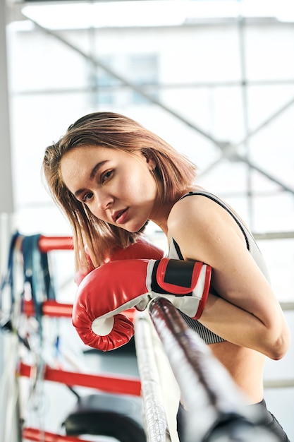 Hübsche junge blonde Sportlerin in roten Boxhandschuhen, die Sie beim Anlehnen an Ringstangen nach dem Training im Fitnessstudio betrachten