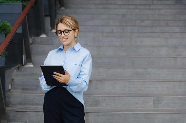 Hübsche junge Blondine mit Brille, blauem Hemd und schwarzer Hose, die im Internet im digitalen Tablet surft, während sie im Freien steht. Konzept moderner Geräte.