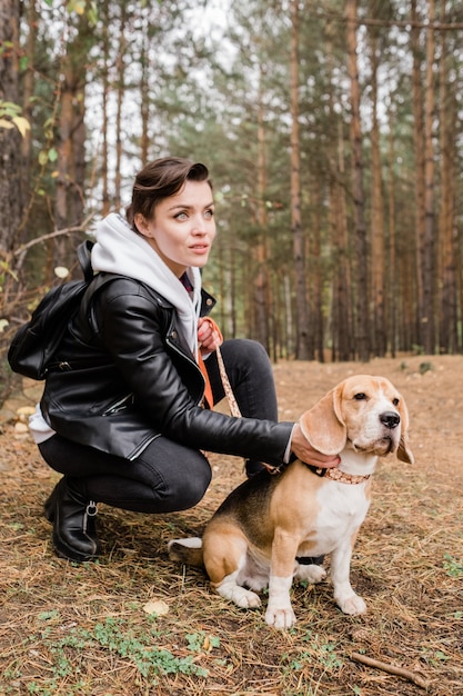 Hübsche junge brünette Frau, die niedlichen Beagle-Welpen kuschelt, während auf Kniebeugen während der Kälte in der natürlichen Umgebung sitzt