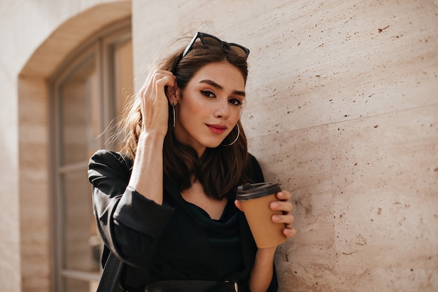 Hübsche junge Brünette mit trendigem Make-up, Brille auf dem Kopf, dunklem Kleid und Jacke, die in der Nähe der beigefarbenen Wand in der Tageslichtstadt steht und eine Tasse Kaffee hält