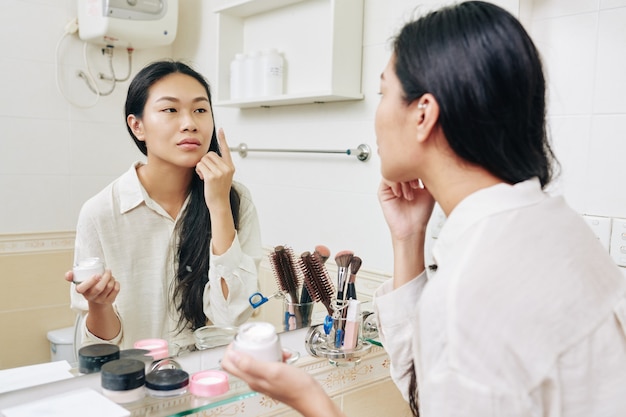 Hübsche junge chinesische Frau, die plumping und feuchtigkeitsspendende Feuchtigkeitscreme vor dem Spiegel im Badezimmer anwendet
