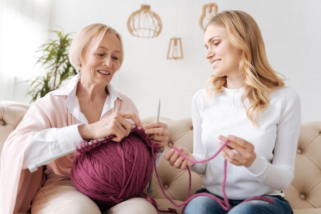 Hübsche junge erwachsene Tochter, die einen Faden für ihre Mutter hält, während sie einen großen Ball von lila Fäden hält und strickt