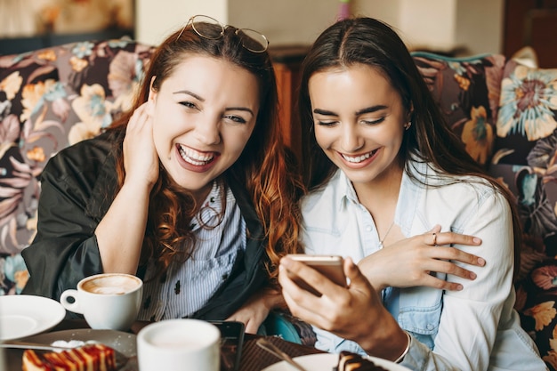 Hübsche junge Frau der Übergröße, die Kamera lachend betrachtet, während ihre Freundin ein lachendes Smartphone beim Sitzen in einem Café betrachtet.