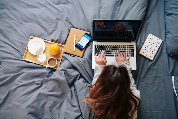 Hübsche junge Frau, die am Laptop im Bett arbeitet, frühstückt und ihr Bankkonto über die Anwendung auf dem Smartphone überprüft