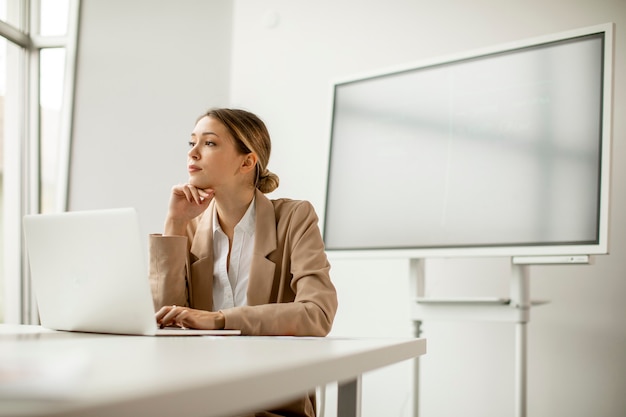 Hübsche junge Frau, die am Laptop im hellen Büro mit großem Bildschirm hinter ihr arbeitet