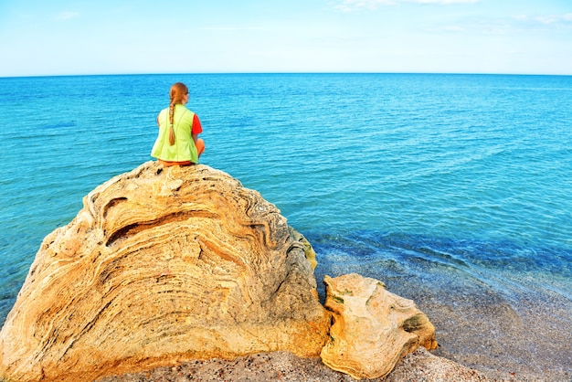 Hübsche junge Frau, die auf einem großen Felsen sitzt und das blaue Meer betrachtet