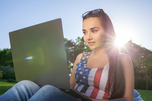 Hübsche junge Frau, die auf Sitzsack sitzt, benutzt Laptop, während sie auf Gras im Park auf der Sonne ruht