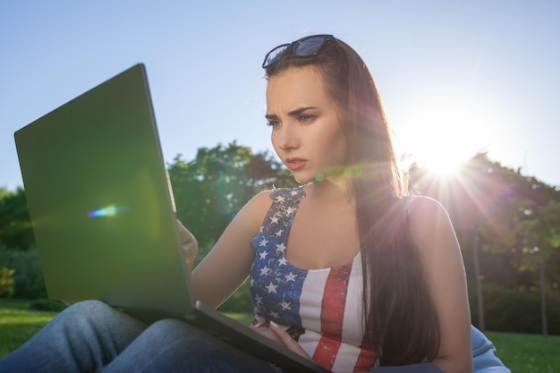 Hübsche junge Frau, die auf Sitzsack sitzt, benutzt Laptop, während sie auf Gras im Park auf der Sonne ruht