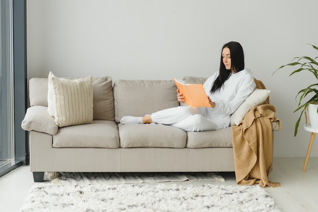 Hübsche junge Frau, die das Lesen eines Buches zu Hause genießt, das auf dem Sofa liegt und im Vergnügen in der Freizeitkleidung lächelt