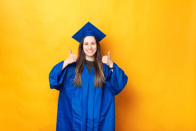 Hübsche junge Frau, die graduierende blaue Kleidung trägt, die Daumen oben zeigt.