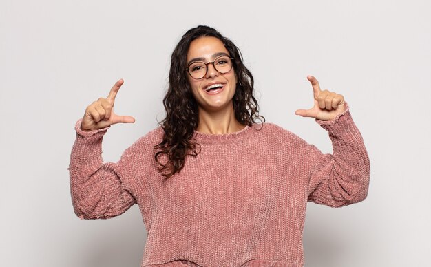 Hübsche junge Frau, die ihr eigenes Lächeln mit beiden Händen einrahmt oder umreißt, positiv und glücklich aussieht, Wellness-Konzept