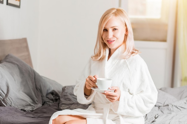 Hübsche junge Frau, die im modernen Hotelzimmer auf dem Bett sitzt, weißen Bademantel trägt, sich nach gesundem Schlaf erfrischt fühlt, guten Service genießt, Kamera beim morgendlichen Teetrinken anschaut.