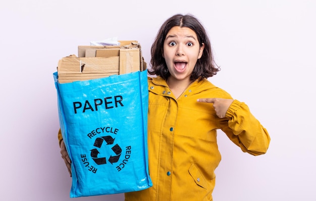 Hübsche junge Frau, die schockiert und überrascht mit weit geöffnetem Mund aussieht und auf sich selbst zeigt. Papier-Recycling-Konzept