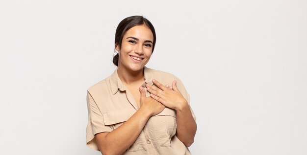 hübsche junge Frau, die sich romantisch, glücklich und verliebt fühlt, fröhlich lächelt und Hände nah am Herzen hält