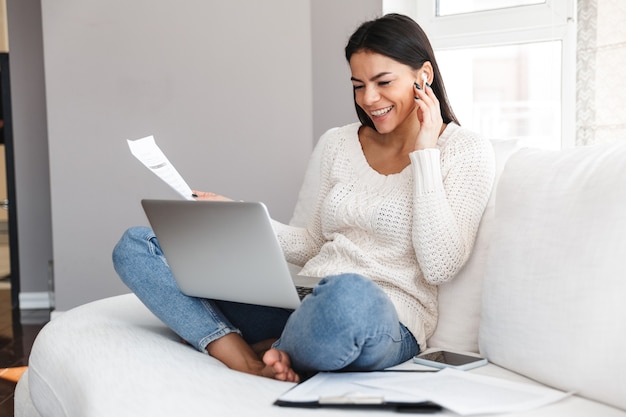 Hübsche junge Frau, die sich zu Hause auf einer Couch entspannt, am Laptop arbeitet und Dokumente hält