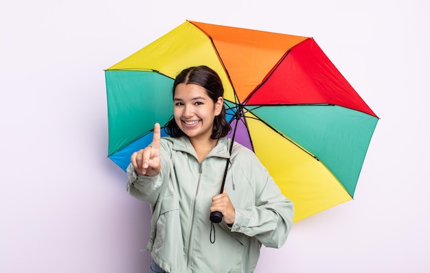 Hübsche junge Frau, die stolz und selbstbewusst lächelt und die Nummer eins macht. Regen- und Regenschirmkonzept