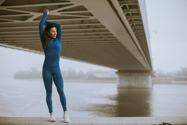 Hübsche junge Frau im blauen Trainingsanzug, die sich am Herbstmorgen vor dem Training am Fluss ausdehnt