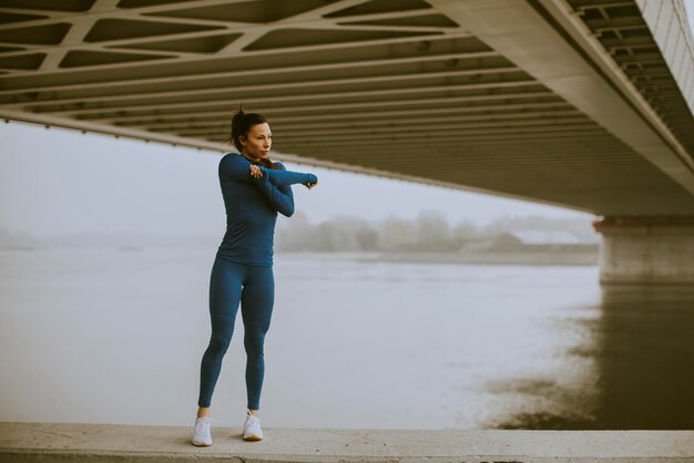 Hübsche junge Frau im blauen Trainingsanzug, die sich am Herbstmorgen vor dem Training am Fluss ausdehnt